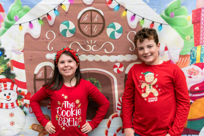 Two children in red holiday shirts at the gingerbread house backdrop