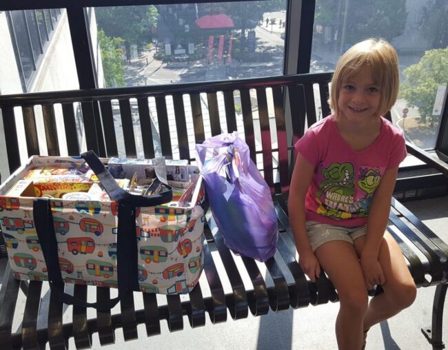 Maci sitting on a bench beside a basket of donated items and a purple bag